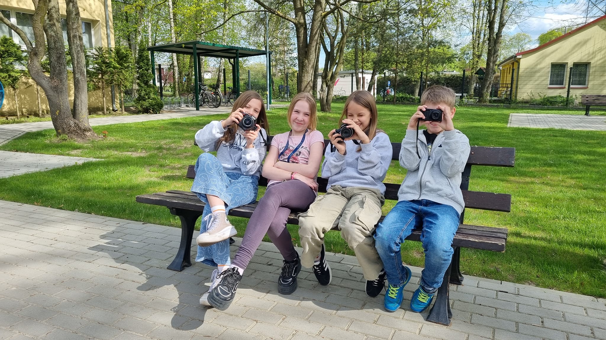 Uczniowie siedzą na ławce przed szkołą i trzymają aparaty fotograficzne 